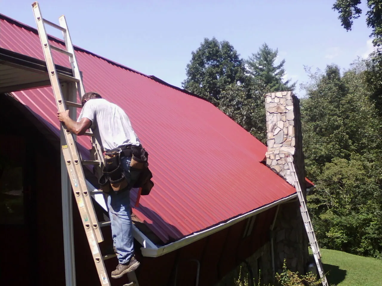 Roofer on a ladder beside a red metal roof and stone chimney — Soffit & Fascia Repair by Roofing Brothers in Waymart